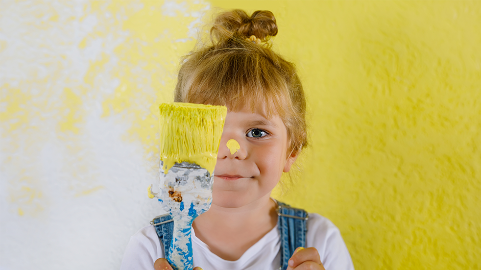 Cute young girl holding up a paintbrush with yellow paint and a half yellow painted wall behind her